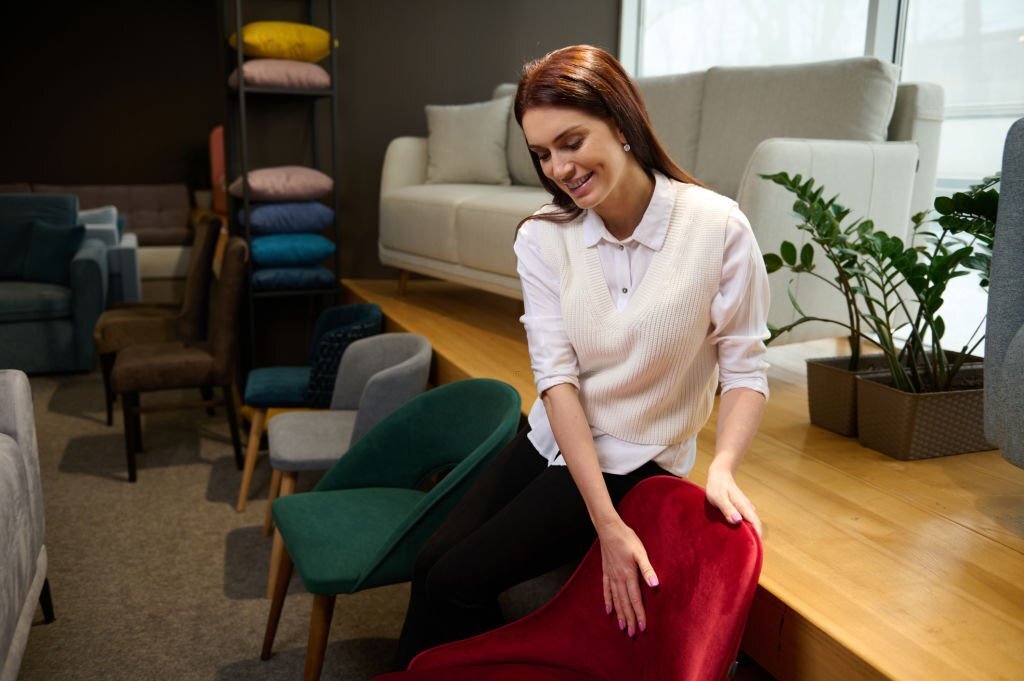 Beautiful pleasant female sales clerk showing the velour armchair in the furniture store showroom. Charming brunette, middle-aged woman choosing for upholstered furniture in the shopping mall